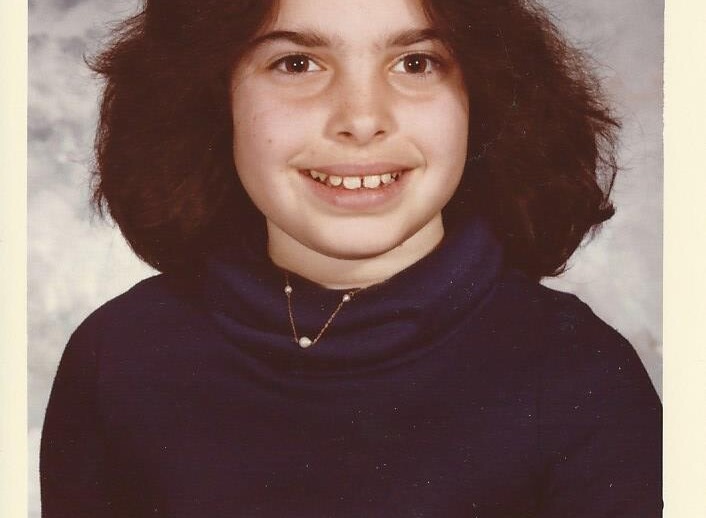 A young person with medium-length dark hair, wearing a navy blue turtleneck, smiles warmly in a portrait-style photo against a gray mottled background, embodying the spirit of SEL activities that promote social and emotional growth.