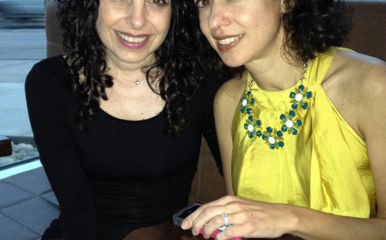 Two women enjoying a meal together at a restaurant. One wears a black top, the other a yellow dress with a beaded necklace. They smile warmly, posing closely with menu and napkin on the table—a picture-perfect moment reflecting the joy of social emotional learning in real life.
