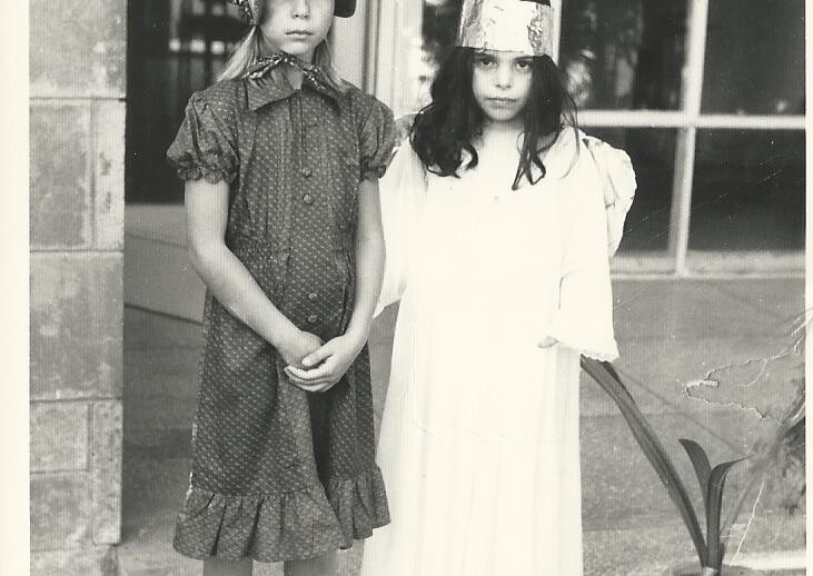 Two children stand outside a building. The one on the left wears a bonnet and a patterned dress, while the one on the right dons a shiny crown and a flowing white dress. This charming duo could be part of fun "get to know you" activities for students in an engaging setting.