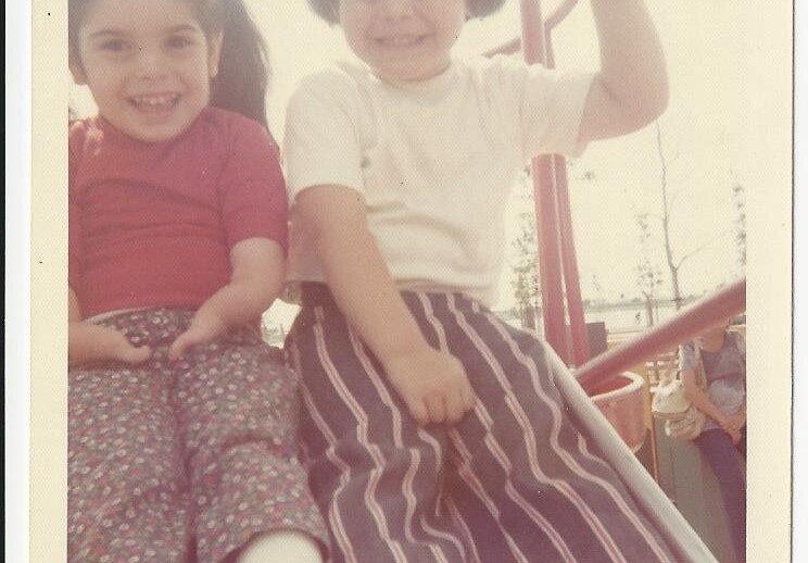 Two children, embodying the spirit of social emotional learning activities, smile joyfully while sitting together on a playground slide. The child on the left wears a red shirt and floral pants, while the one on the right sports a white shirt and striped pants, both radiating playful happiness.