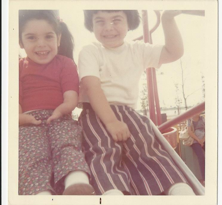 Two children, embodying the spirit of social emotional learning activities, smile joyfully while sitting together on a playground slide. The child on the left wears a red shirt and floral pants, while the one on the right sports a white shirt and striped pants, both radiating playful happiness.