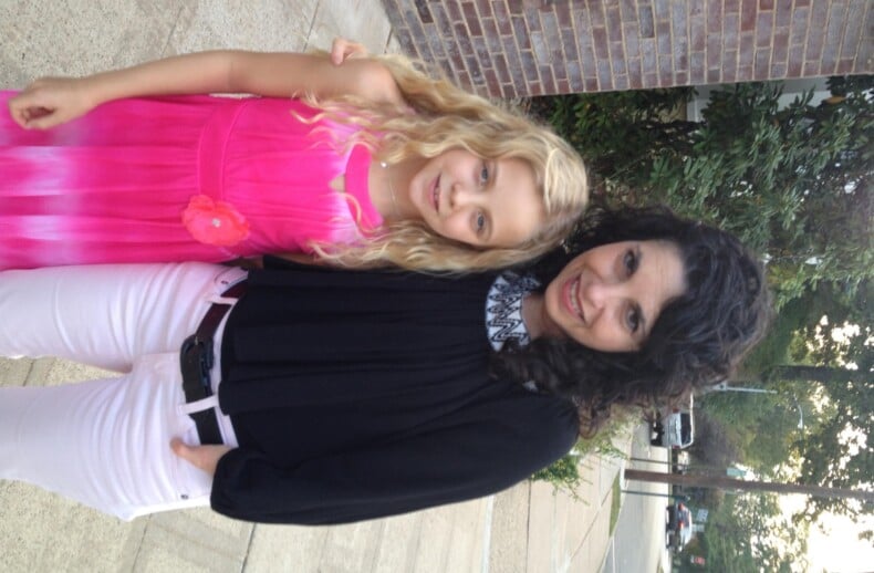 A woman with curly dark hair stands next to a young girl with long blonde hair, exemplifying the essence of diversity programs. The woman wears a black top and white pants, while the girl sports a pink dress. They are outdoors on a sidewalk, with trees and a brick building in the background.
