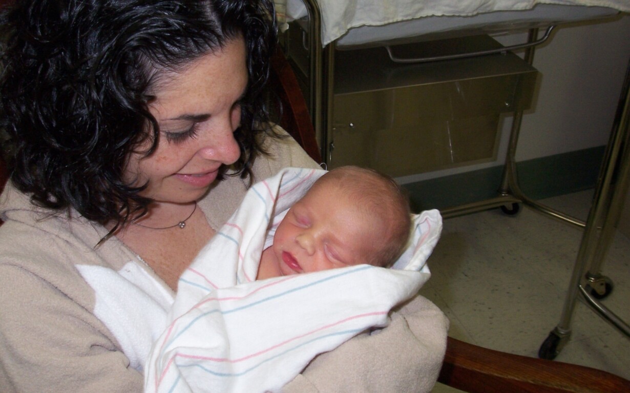 A woman with curly dark hair sits in a hospital chair, smiling gently as she cradles a newborn baby wrapped in a white blanket. The scene, rich with emotion, is reminiscent of the warmth fostered by SEL activities in nurturing environments like hospitals.
