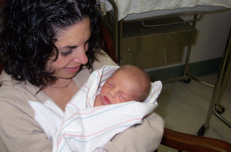 A woman with curly dark hair sits in a hospital chair, smiling gently as she cradles a newborn baby wrapped in a white blanket. The scene, rich with emotion, is reminiscent of the warmth fostered by SEL activities in nurturing environments like hospitals.