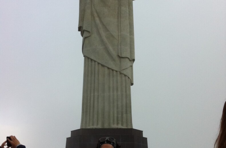 A woman poses with arms outstretched in front of the large Christ the Redeemer statue in Brazil, embracing a moment that evokes SEL principles. Several other tourists, drawn into this social-emotional exploration, gather around the base. The sky is cloudy in the background.