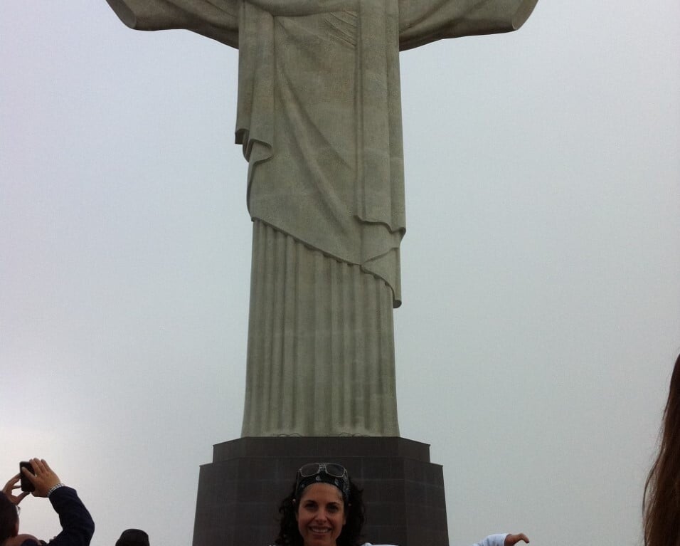 A woman poses with arms outstretched in front of the large Christ the Redeemer statue in Brazil, embracing a moment that evokes SEL principles. Several other tourists, drawn into this social-emotional exploration, gather around the base. The sky is cloudy in the background.