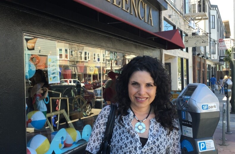 A person with curly hair and a patterned blouse stands next to a city street parking meter. Behind them, a storefront with "826 Valencia" on the awning hints at creativity, much like SEL activities fostering growth and understanding in diverse urban communities.