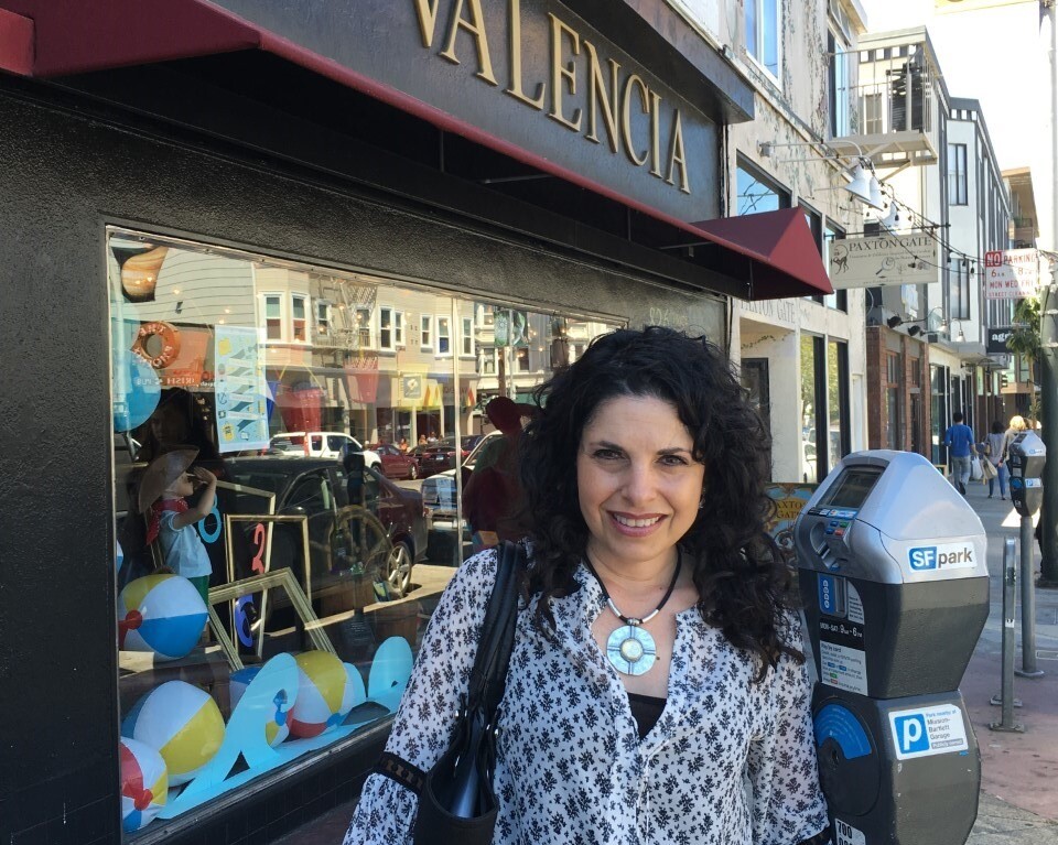 A person with curly hair and a patterned blouse stands next to a city street parking meter. Behind them, a storefront with "826 Valencia" on the awning hints at creativity, much like SEL activities fostering growth and understanding in diverse urban communities.