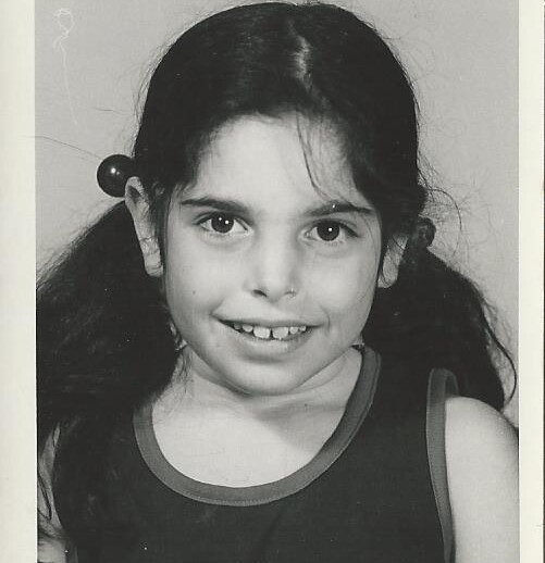 A black and white portrait of a young girl with long dark hair in pigtails and a sleeveless top. She is smiling at the camera, embodying the joy often found in social emotional learning activities.