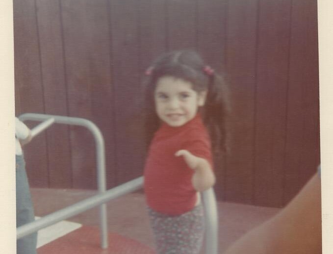 A young child with long dark hair in pigtails stands on a playground merry-go-round, wearing a red shirt and patterned pants. A wooden fence is in the background as part of their SEL activities. The child is smiling at the camera.