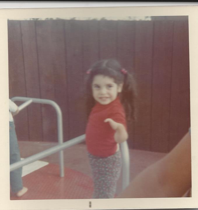A young child with long hair in pigtails, wearing a red shirt and patterned pants, stands on a playground merry-go-round. This joyful scene highlights the impact of social emotional learning activities, fostering connection beyond the brown wooden fence in the background.
