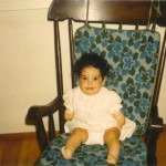 A baby with curly hair sits happily on a wooden rocking chair with a floral cushion, embodying the joy and comfort often emphasized in social emotional learning curricula. Wearing a light-colored outfit, the little one brightens the room's wooden floor and plain walls.