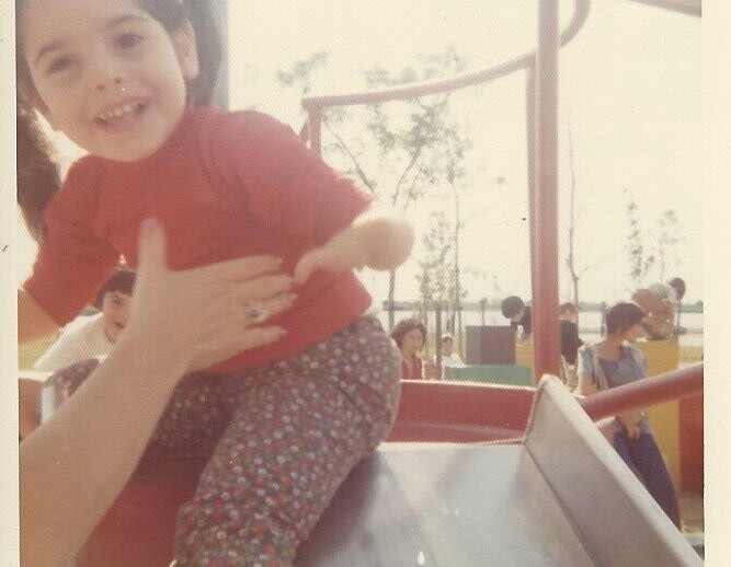 A young child in a red shirt and patterned pants smiles while sliding down a playground slide, an example of social emotional learning activities. An adult's arms are extended to support the child. In the background, other children and trees are visible, fostering community bonds.