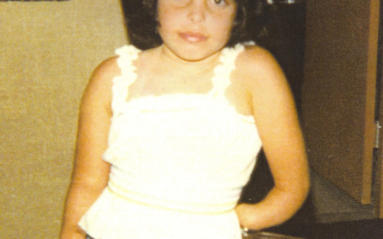 A young girl with dark hair stands in a room, wearing a white sleeveless top and denim shorts. She leans slightly against a chair with one hand in her pocket, embodying the confidence taught through social emotional learning activities. The background includes a door and a closet.