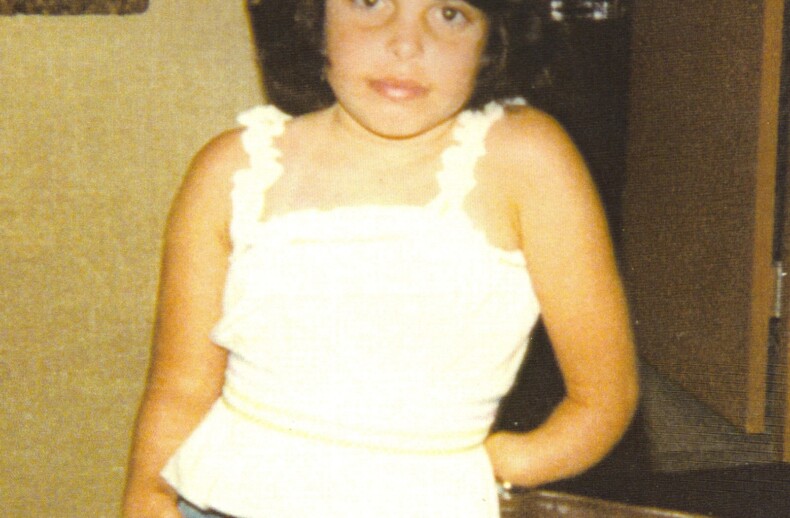 A young girl with dark hair stands in a room, wearing a white sleeveless top and denim shorts. She leans slightly against a chair with one hand in her pocket, embodying the confidence taught through social emotional learning activities. The background includes a door and a closet.