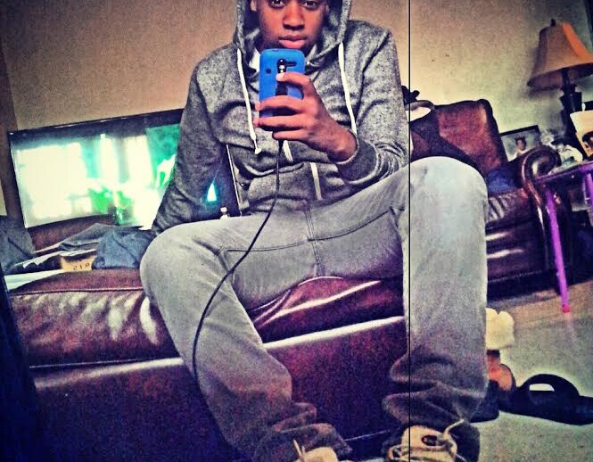 A person in a gray hoodie and sweatpants takes a mirror selfie while sitting on a brown leather couch, holding a blue phone. The casually furnished room reflects an everyday backdrop, much like those often depicted in social emotional learning activities, with a TV, lamp, and clothes scattered around.
