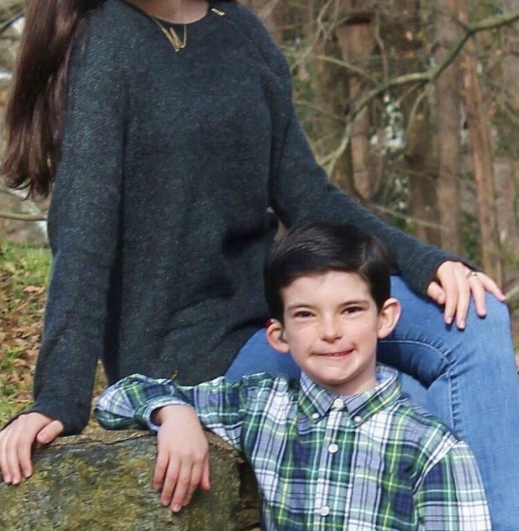 A girl and a young boy smile at the camera while sitting on a stone wall, embodying the spirit of SEL activities. The girl is wearing a dark sweater and jeans, and the boy is in a plaid shirt. The background features trees and foliage, creating an inviting atmosphere for growth and connection.