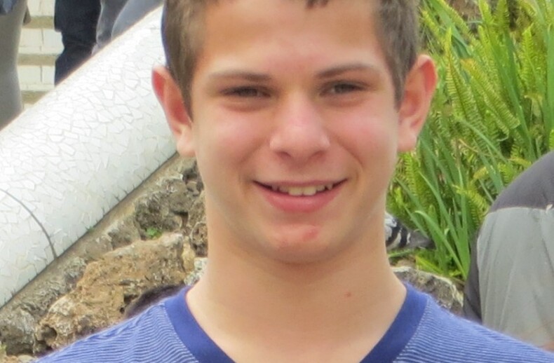 A young man with short brown hair smiles while standing outdoors, embodying the spirit of social emotional learning curriculum. He is wearing a blue-striped shirt, and there is greenery and a stairway in the background.