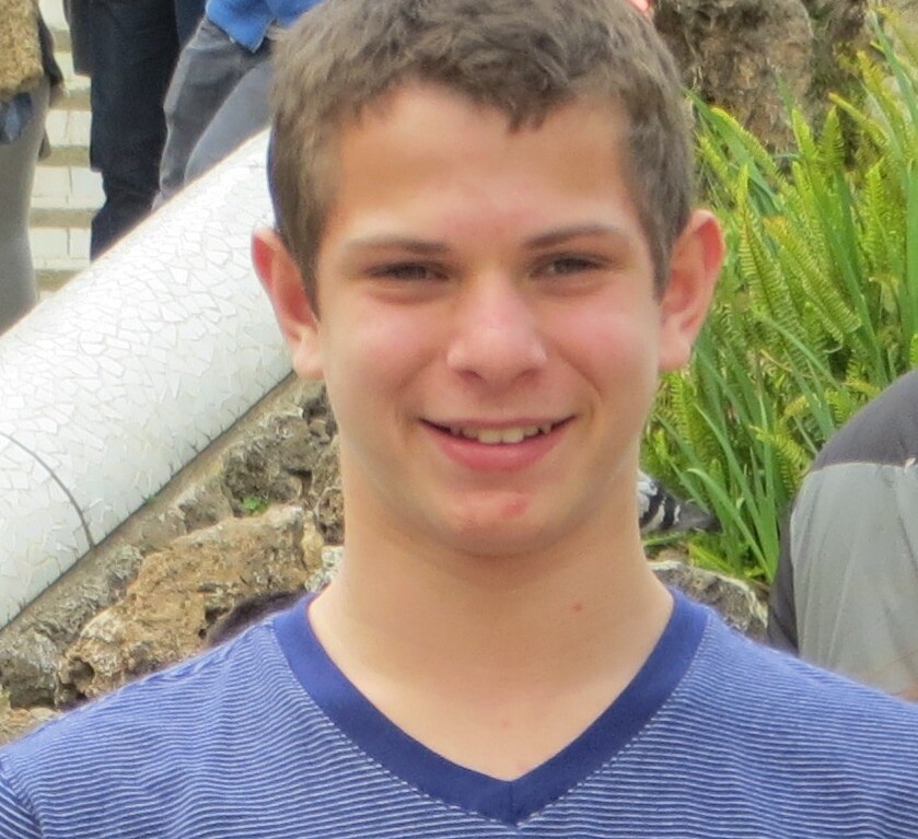 A young man with short brown hair smiles while standing outdoors, embodying the spirit of social emotional learning curriculum. He is wearing a blue-striped shirt, and there is greenery and a stairway in the background.
