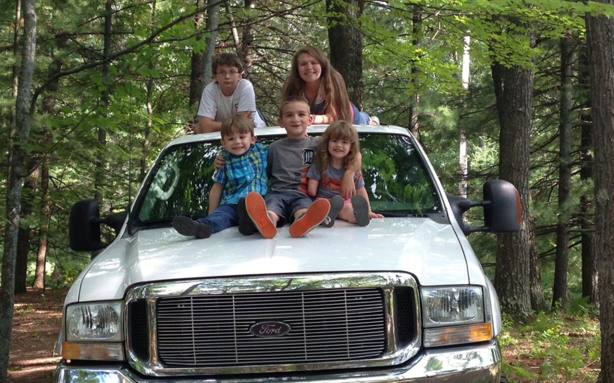 A group of five children sit on the hood of a white Ford truck parked in a forest, engaging in get-to-know-you activities. The kids are smiling and dressed casually, with trees and greenery providing a natural backdrop that mirrors their growing camaraderie.
