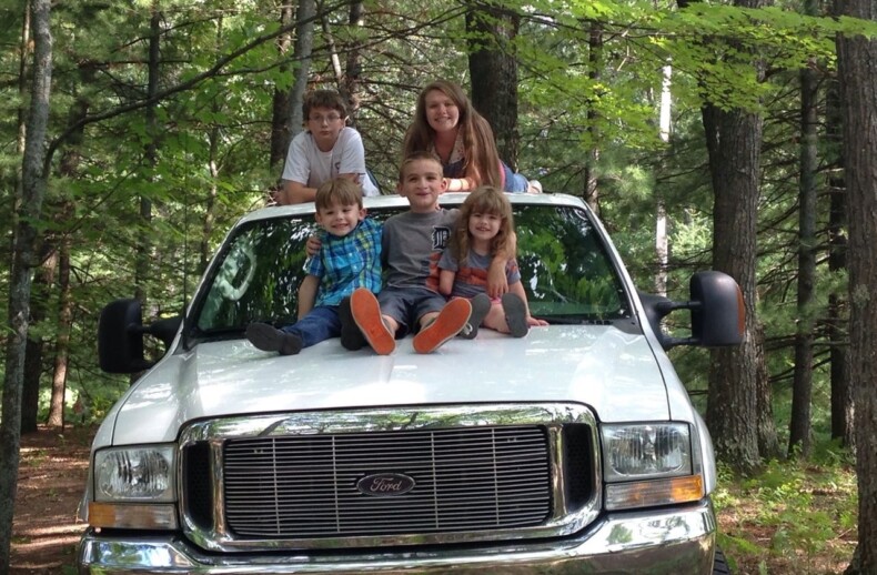 A group of five children sit on the hood of a white Ford truck parked in a forest, engaging in get-to-know-you activities. The kids are smiling and dressed casually, with trees and greenery providing a natural backdrop that mirrors their growing camaraderie.
