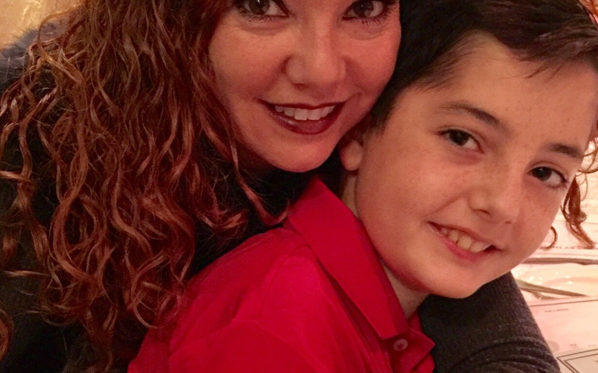 A woman with curly red hair embraces a young boy with dark hair. They are both smiling, seated at a table covered with a white tablecloth in a warmly lit room, reflecting the joy found in social emotional learning activities.