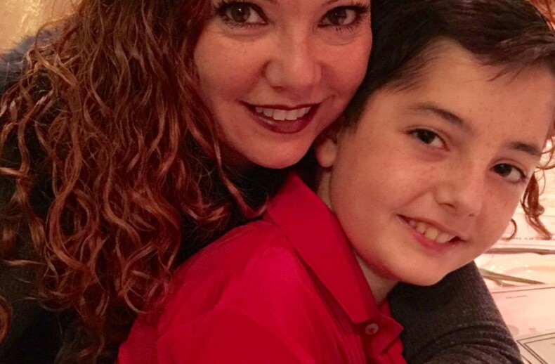 A woman with curly red hair embraces a young boy with dark hair. They are both smiling, seated at a table covered with a white tablecloth in a warmly lit room, reflecting the joy found in social emotional learning activities.