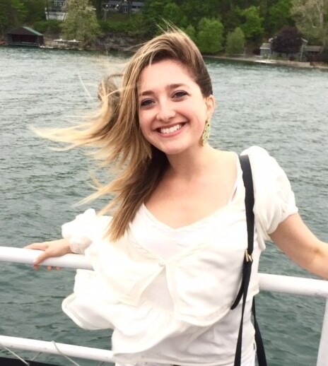 A smiling woman with long blonde hair stands on a boat, embracing the calming influence of nature as part of her social emotional learning activities. She holds onto the railing, her white outfit bright against the backdrop of water and distant trees, while her hair dances in the wind.
