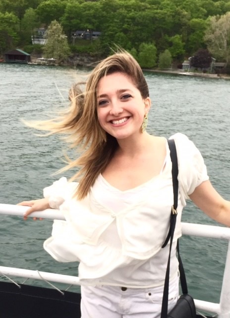 A smiling woman with long blonde hair stands on a boat, embracing the calming influence of nature as part of her social emotional learning activities. She holds onto the railing, her white outfit bright against the backdrop of water and distant trees, while her hair dances in the wind.