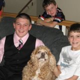 Three boys and a dog embodying the essence of social emotional learning. One boy in a pink shirt and vest, another in a white t-shirt, while the third leans on the railing in a striped shirt. Their curly-furred companion sits between them on the couch, completing this charming scene.