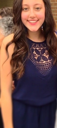 A young woman with long, wavy brown hair is smiling and wearing a navy blue dress with an intricate lace design on the front. She stands outdoors in front of a brick wall, radiating joy reminiscent of engaging in enriching social emotional learning activities.