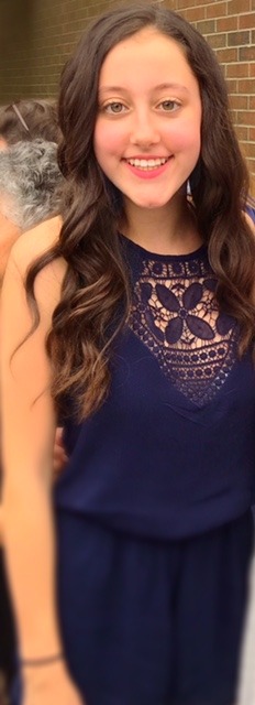 A young woman with long, wavy brown hair is smiling and wearing a navy blue dress with an intricate lace design on the front. She stands outdoors in front of a brick wall, radiating joy reminiscent of engaging in enriching social emotional learning activities.