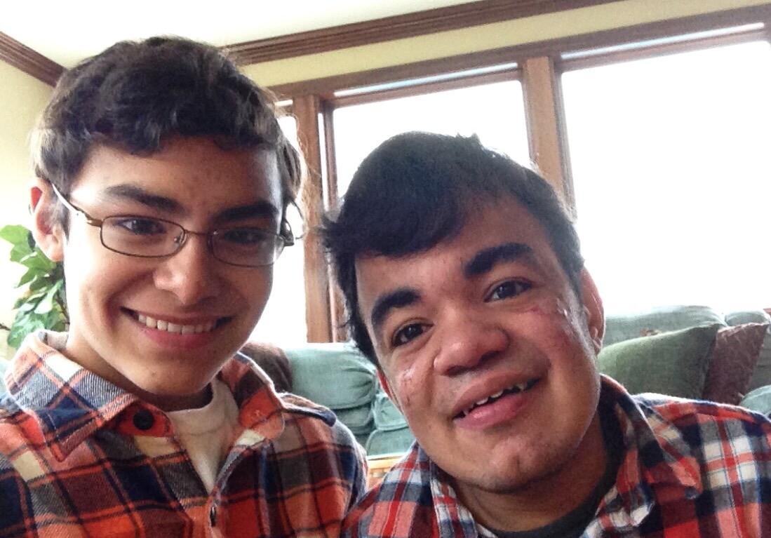 Two people smiling at the camera, both wearing glasses and plaid shirts. They are indoors with a large window behind them, inviting natural light as they engage in social emotional learning activities.