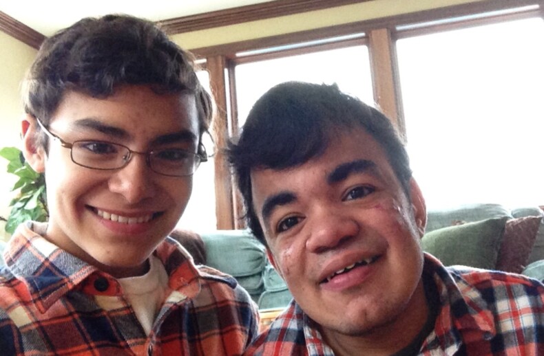 Two people smiling at the camera, both wearing glasses and plaid shirts. They are indoors with a large window behind them, inviting natural light as they engage in social emotional learning activities.