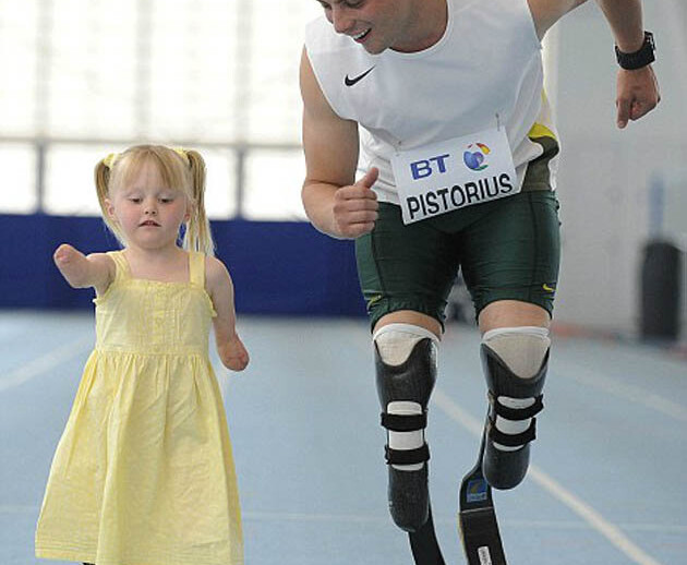 A man with prosthetic legs and a young girl, also using prosthetics, run side by side on an indoor track. Part of a dynamic SEL curriculum, the girl's yellow dress flutters as they smile joyfully, embodying the spirit of inclusive diversity programs through their shared activity.