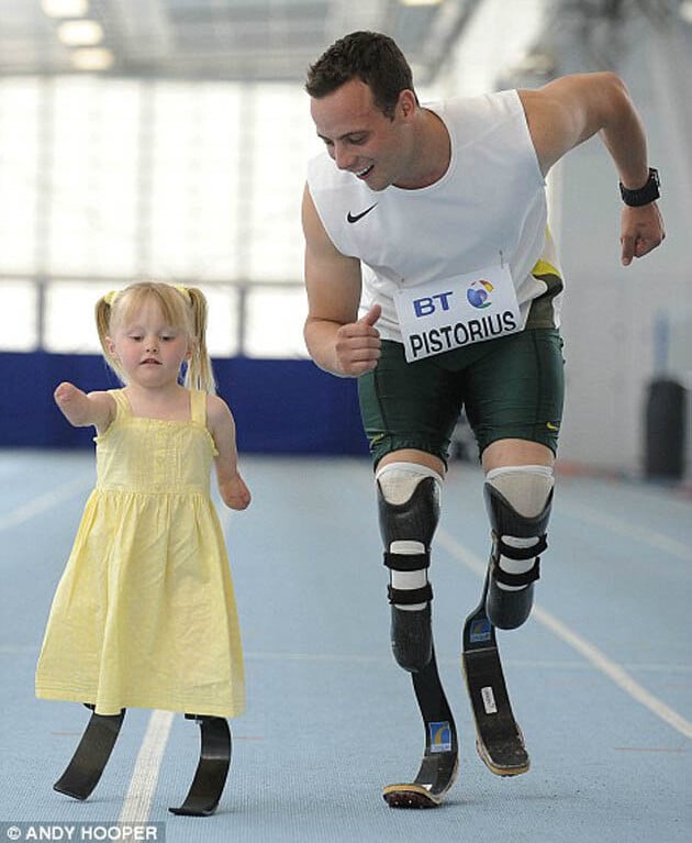 A man with prosthetic legs and a young girl, also using prosthetics, run side by side on an indoor track. Part of a dynamic SEL curriculum, the girl's yellow dress flutters as they smile joyfully, embodying the spirit of inclusive diversity programs through their shared activity.
