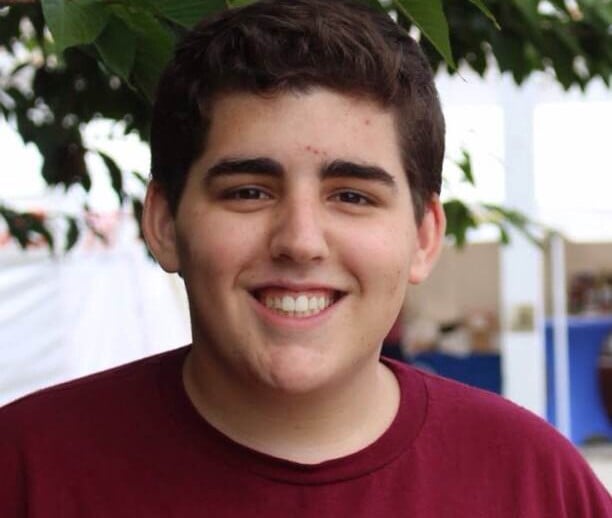 A person with short dark hair is smiling at the camera, wearing a maroon shirt. They stand outdoors under tree branches and lush green leaves, embodying the spirit of diversity programs. The blurred background features a white structure, adding depth to this vibrant scene.
