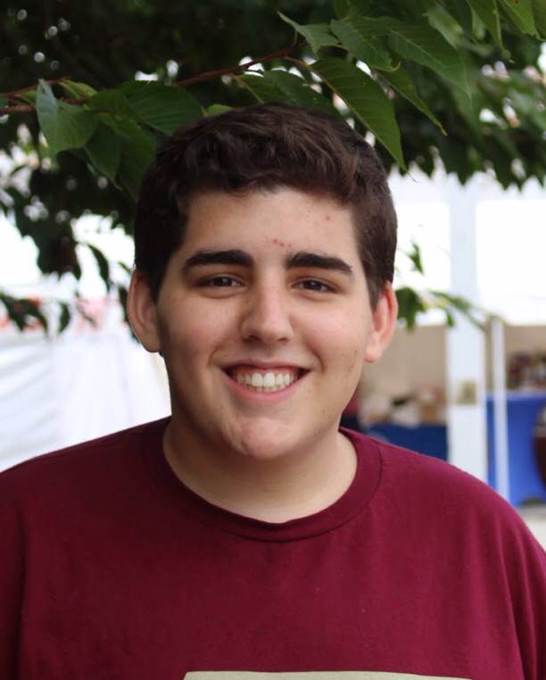 A person with short dark hair is smiling at the camera, wearing a maroon shirt. They stand outdoors under tree branches and lush green leaves, embodying the spirit of diversity programs. The blurred background features a white structure, adding depth to this vibrant scene.