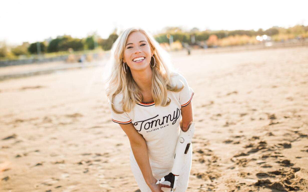 A woman with long blonde hair smiles brightly on a sandy beach, embodying resilience like the insights fostered by SEL activities. She wears a white shirt with "Tommy" written on it and has a prosthetic arm. The background captures greenery and the sunlit horizon.