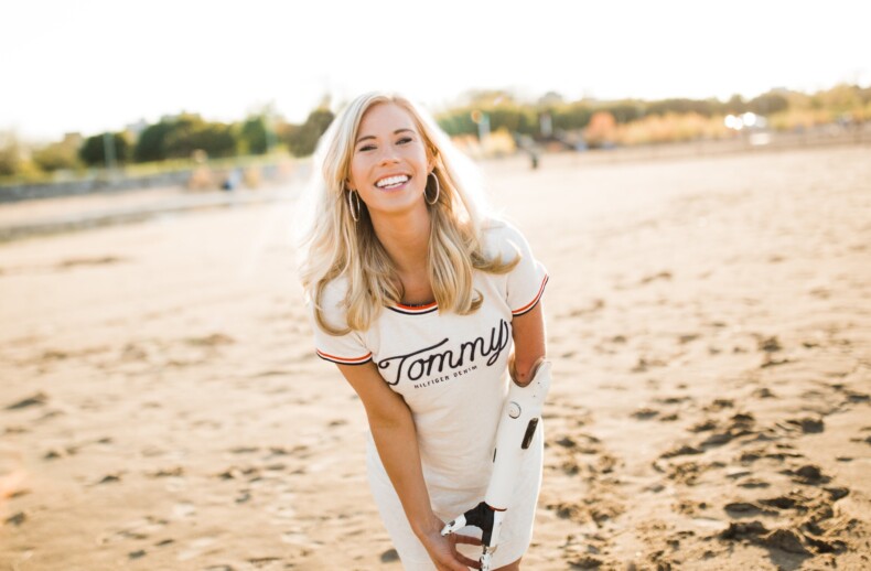 A woman with long blonde hair smiles brightly on a sandy beach, embodying resilience like the insights fostered by SEL activities. She wears a white shirt with "Tommy" written on it and has a prosthetic arm. The background captures greenery and the sunlit horizon.