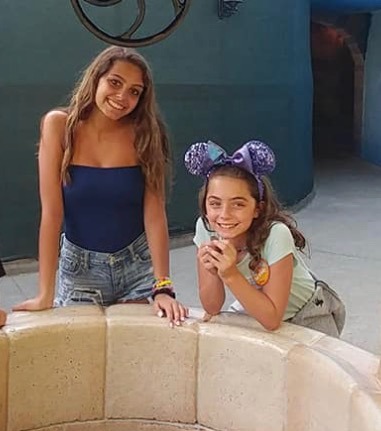 Two smiling girls lean on a stone ledge. One wears a blue top and denim shorts; the other sports a light top with purple Mickey Mouse ears headband. In a playful mood, they seem like they're enjoying get-to-know-you activities in an outdoor setting.