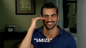 A man with dark hair and a beard is smiling while signaling with his hand near his face. The word "SMIZE" is written in bold white letters at the bottom. He is wearing a blue shirt and sitting indoors, embodying the warmth often cultivated in SEL activities.