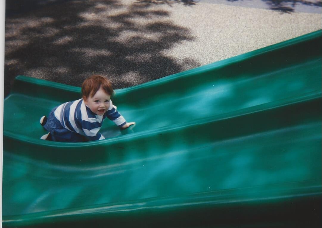 A young child in a striped shirt, engaging in social emotional learning activities, crawls up a green slide, smiling. The background features dappled sunlight on the ground.