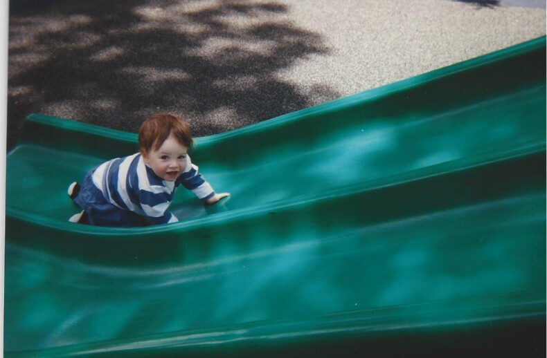 A young child in a striped shirt, engaging in social emotional learning activities, crawls up a green slide, smiling. The background features dappled sunlight on the ground.