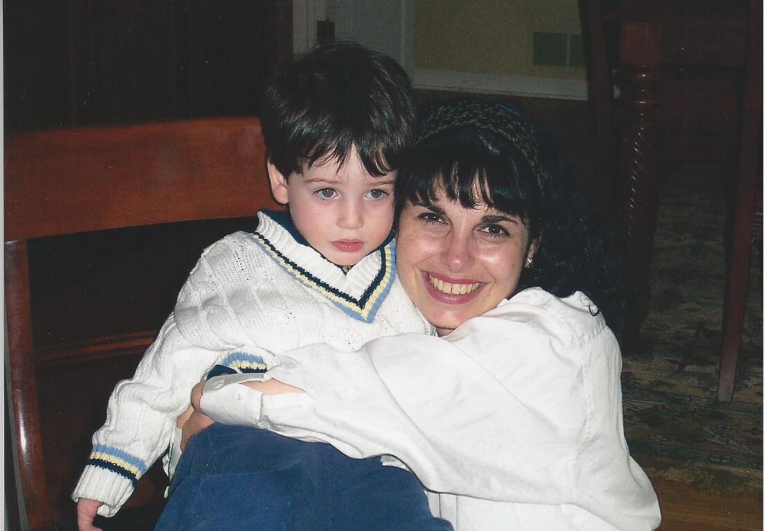 A woman with dark hair is smiling and hugging a young boy with brown hair, showcasing the warmth fostered by social emotional learning activities. The boy is wearing a white sweater with blue and yellow trim, and blue pants. They are sitting on a wooden chair indoors.