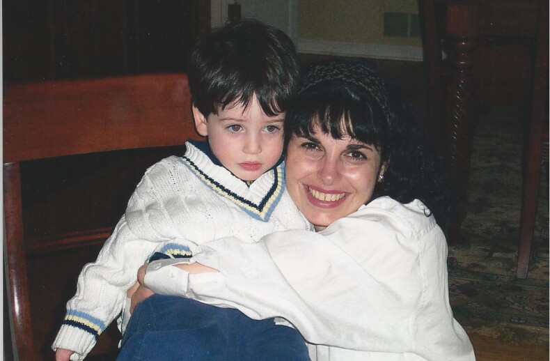A woman with dark hair is smiling and hugging a young boy with brown hair, showcasing the warmth fostered by social emotional learning activities. The boy is wearing a white sweater with blue and yellow trim, and blue pants. They are sitting on a wooden chair indoors.