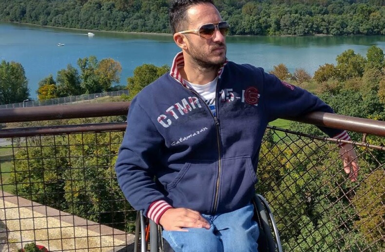 A man in a wheelchair wearing sunglasses and a navy blue hoodie poses in front of a scenic view with a river, trees, and city skyline. It's a sunny day with a partly cloudy sky—a perfect setting for SEL activities that connect with nature and spark meaningful interactions.