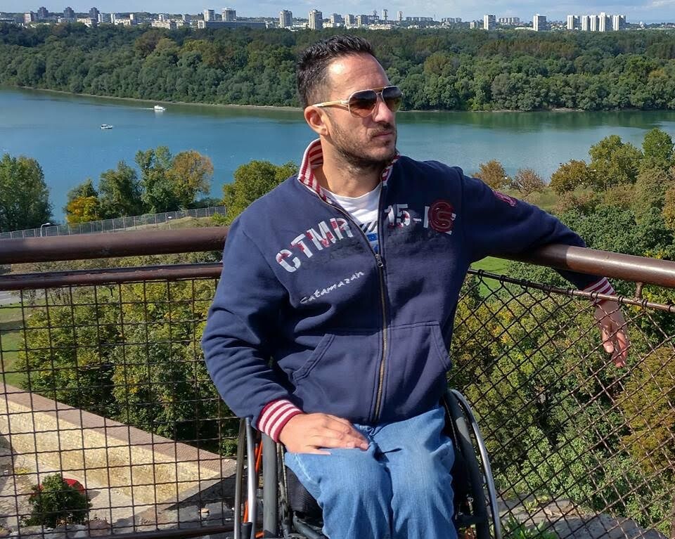 A man in a wheelchair wearing sunglasses and a navy blue hoodie poses in front of a scenic view with a river, trees, and city skyline. It's a sunny day with a partly cloudy sky—a perfect setting for SEL activities that connect with nature and spark meaningful interactions.