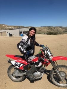 A person wearing motocross gear sits on a red dirt bike numbered 94, smiling under the clear blue sky. With a building and rolling hills in the background, it's an exhilarating scene that sparks social emotional learning activities by embracing adventure and confidence.