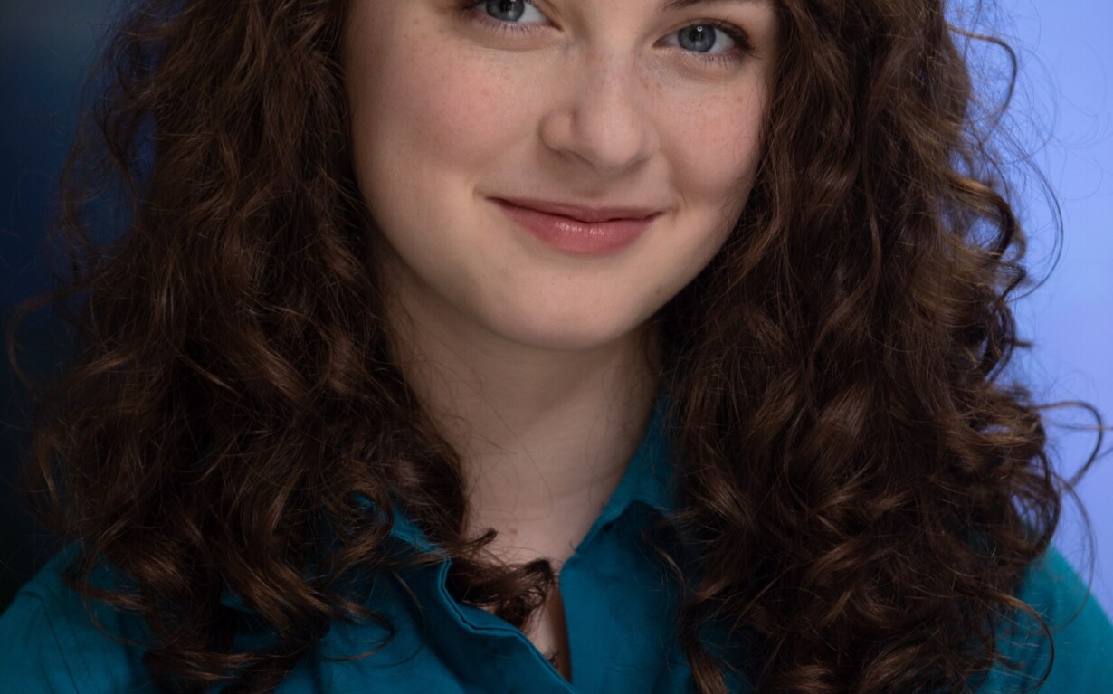 A person with long, curly brown hair smiles gently at the camera. They wear a teal shirt against a blurred blue background, reflecting the welcoming and inclusive spirit of diversity programs while presenting a professional and friendly appearance.